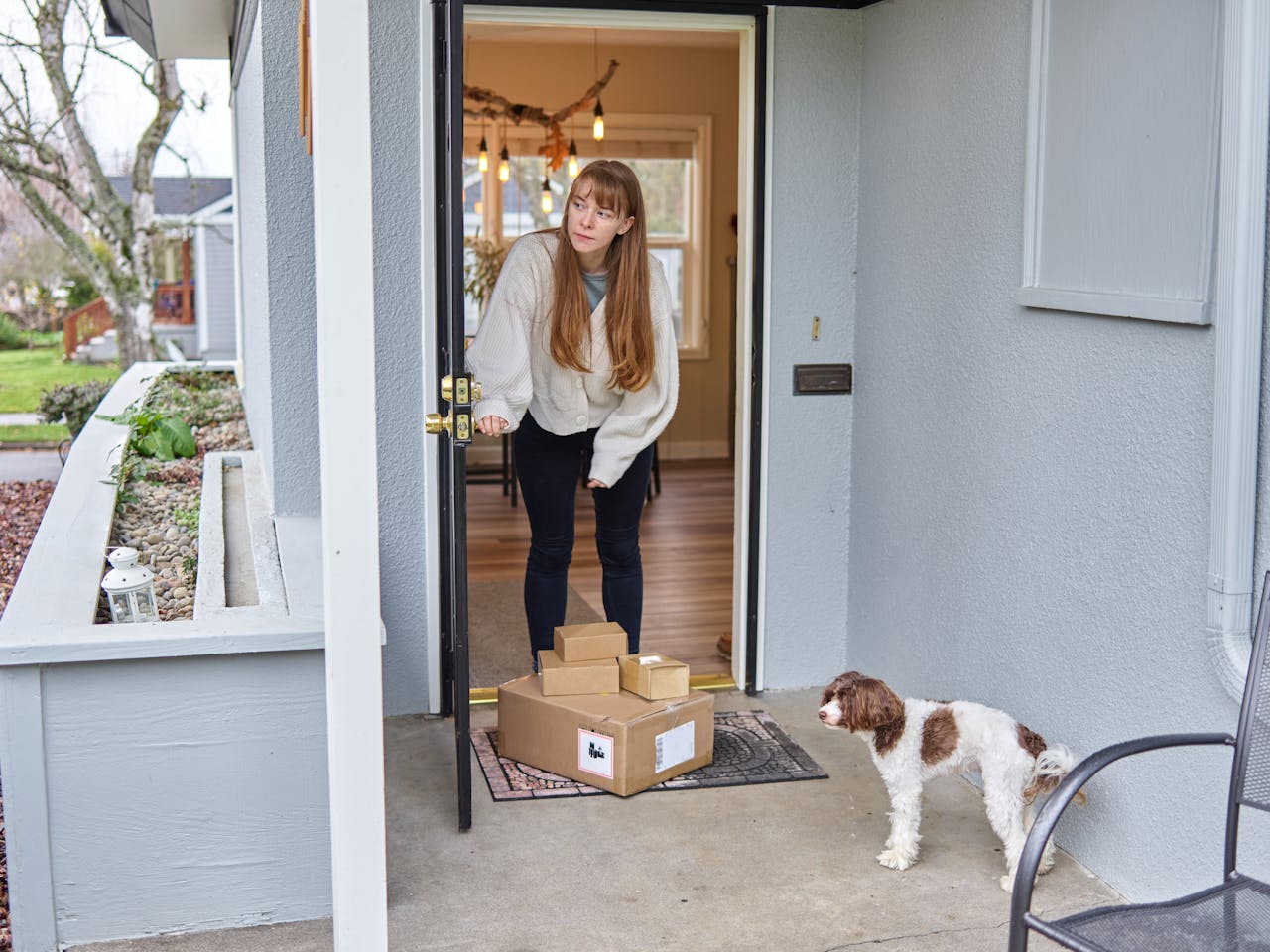 hero-img-01 Woman opens front door to receive packages delivered on porch. Dog observes.